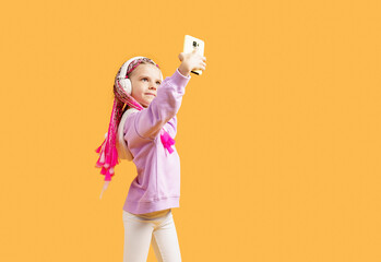 Pretty modern girl with pink dreadlocks in headphones smiling and making selfie on smartphone isolated over yellow.