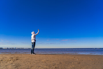 A young woman stands on the beach and films herself on a mobile phone from afar. Space for text.