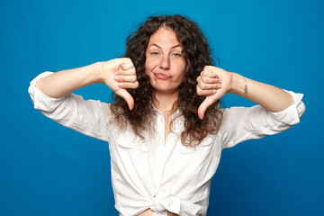 Curly  haired female expresses dislike with body language sign , keeps thumbs down and frowns face, has displeased angry expression, sun-tanned skin, curves lips, isolated over blue wall background. © Yulia