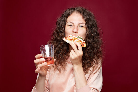 Close up indoor portrait of festive joyful female, biting pizza slice with passion and hunger, holding plastic glass with lemonade or sparkling wine, has curly hairstyle, posing on crimson studio wall