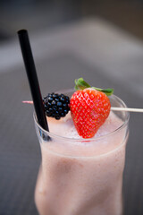 Vertical closeup of a transparent glass with fresh milk shake with strawberry and blackberry decorated with berries on a stick and black cocktail tube. Served in outdoor bar on grey table. smoothie