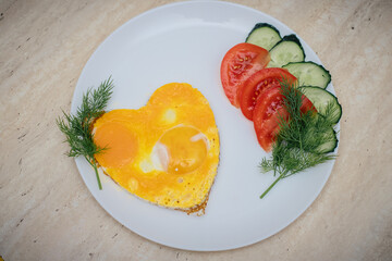 fried eggs in the form of a heart lie on a white plate, next to chopped dill, tomatoes and cucumbers