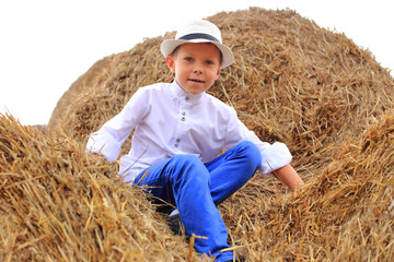 Naklejka na ściany i meble A cute boy is high under the sky on a haystack in the hay season.