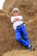 Naklejka na ściany i meble A cute boy is high under the sky on a haystack in the hay season.