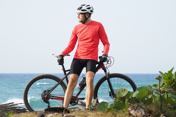 Fitness man with a bicycle stands on the ocean shore and looks into the distance on a sunny day. Athletic healthy people active lifestyle during pandemic coronavirus..