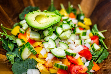 Chopped sliced vegetables in wooden bowl macro closeup with fresh vegan salad with baby kale, tomatoes, bell peppers and avocado half