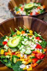Chopped sliced vegetables in wooden bowls closeup with fresh vegan salad with baby kale, tomatoes, bell peppers and avocado half