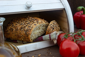 Fresh baked bread with seeds (sesame, sunflower seeds, linen) in white wooden bread box. Breadbox with cutted bread, tomatoes, olive oil in glass bottle and red bell peppers on the wooden background. - obrazy, fototapety, plakaty