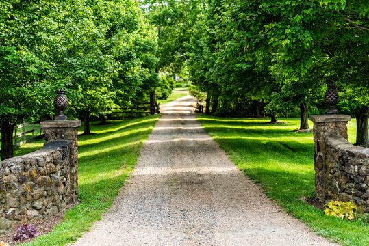 Gated open entrance with road driveway in rural countryside in Virginia estate with stone fence and gravel dirt path street with green lush trees in summer