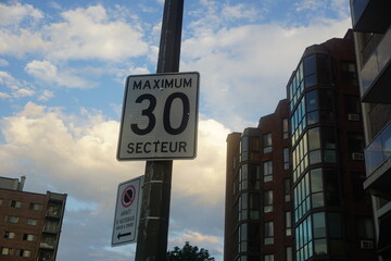 A traffic sign speed limit of 30 Km/hr in a sector with a background of a cloudy blue sky. No stop sign to the left says a bus stop from 5:30 am to 8:00 pm.