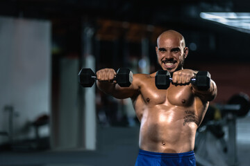 Fototapeta na wymiar Portrait of young exhausted strong man lifting two dumbbells while exercising in gym