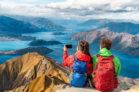 Hikers looking at view on mountain top summit on hiking travel vacation - couple taking photo with phone. Wanderlust adventure people relaxing. Summit of famous hike to Roys Peak, New Zealand.