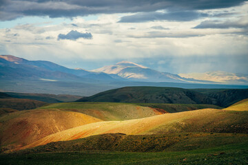 Awesome vast landscape with vivid multicolor mountains at sunset. Scenic view to colorful hills and big mountains under cloudy sky. Picturesque sunny scenery with awesome mountains in rainy weather.