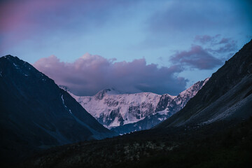 Amazing landscape of sunset with pink snowy mountains and lilac clouds. Atmospheric highland scenery with great mountain silhouettes under purple dawn sky. Awesome view to violet snow peak in clouds.