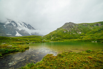 Vivid green landscape with raindrops on mountain lake among low clouds. Rain drops on lake water. Colorful scenery of highlands in rainy weather. Circles on green water. Droplets on alpine lake.