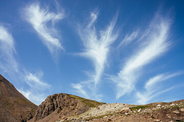 Scenic mountain landscape with beautiful cirrus clouds in clear blue sky over rocks in sunlight. Colorful highland scenery with spindrift clouds in blue clear sky above green brown rocky mountains.