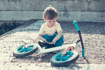 A handsome boy of 2 years old is looking at a bicycle lying at his feet. Child and bike without pedals. Sorry kid. Close-up, toned