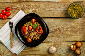 On a wooden table is a black plate of boiled mung bean with vegetables and mushrooms on a napkin and a fork. Copy space.