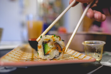 Close up chopstick holding a vegan sushi roll with seitan meat, avocado, green onion and carrot and in a plate with orange and white leaf pattern design