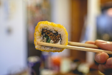 Close up chopstick and fingers holding a vegan sushi roll with seitan meat, vegan cream cheese, green onion and fried tempura 