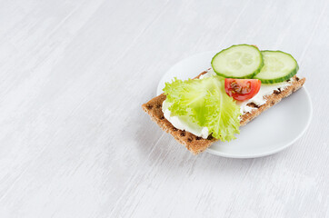 Organic season sandwiches on dry brown grain crispbread with fresh sliced vegetables - green salad, cucumber, tomato, cream cheese on white wooden table, closeup, copy space.
