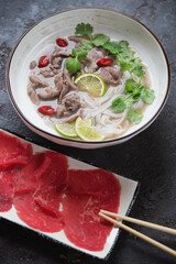 Pho bo soup served with raw beef meat slices, vertical shot on a brown stone surface, selective focus