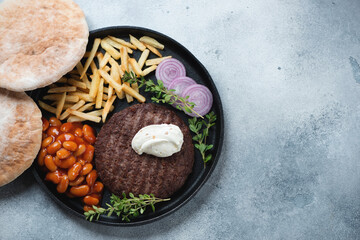 Grilled balkan pljeskavica served with fries, baked beans and pitas on a cast-iron tray, top view on a light-blue stone background, copyspace