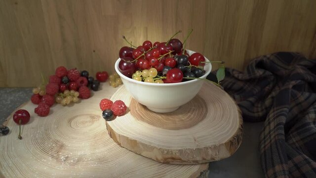 Raspberries, cherries, white and red currants, black currants in a white bowl on wooden pieces of tree trunk. Sun glare moves across the background