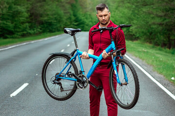 Obraz premium Athletic Man in a tracksuit holds a bicycle on his shoulders while standing on the road in the forest. The concept of a healthy lifestyle, cardio training. Copyspace.