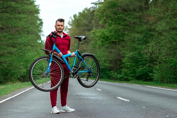 Athletic Man in a tracksuit holds a bicycle on his shoulders while standing on the road in the forest. The concept of a healthy lifestyle, cardio training. Copyspace.