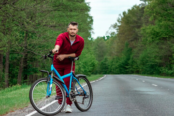 Obraz premium A man in a tracksuit stands next to a bicycle on a road in the forest. The concept of a healthy lifestyle, cardio training. Copyspace.