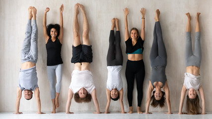 Fototapeta na wymiar Happy young multiracial women in activewear standing near wall with raised arms while sportive mixed race people performing handstands, having fun together before intensive workout in gym indoors.