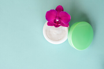 Fototapeta na wymiar Cream in white jar with green cap on mint background with beautiful bright magenta orchid flowers. Soft cream with orchid extract for moisturizing skin. Eco cosmetic product, top view
