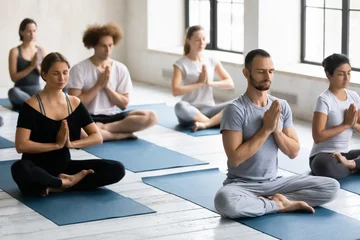 Tuinposter Young motivated multiracial people in activewear sitting on floor mats in lotus position with folded in namaste hands, feeling thankful after workout or enjoying meditating with closed eyes indoors. © fizkes