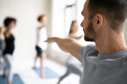 Close up back rear view young focused professional fit male trainer giving master class to group of students, showing warrior second virabhadrasana II position to motivated people in modern studio.