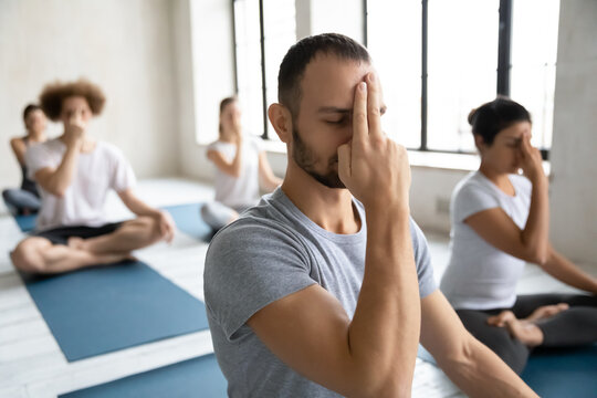 Head shot young mindful male trainer leading yoga group class, showing Alternate Nostril Breathing to motivated diverse students, practicing nadi shodhana pranayama together finishing workout indoors.