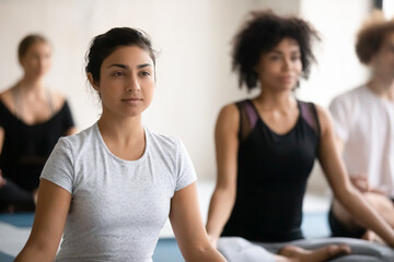 Focus on peaceful happy young indian ethnicity woman beginner, enjoying yoga master class with diverse people students, learning meditation or relaxing muscles after intensive workout indoors.