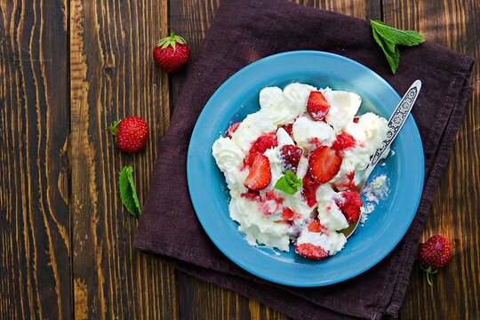 Dessert known as eton mess, crispy meringues, whipped cream and fresh strawberries on a blue plate on a wooden background.