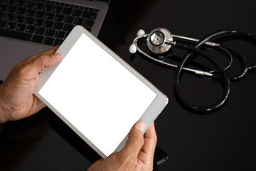 Mockup hands hold digital tablet with blank white screen and medical stethoscope with laptop computer on desk  isolated on dark background. Medical technology, medic tech concept. Top view. 