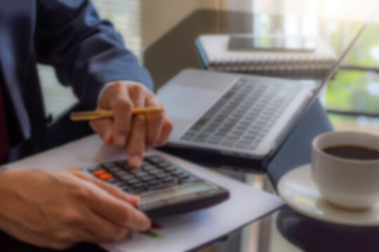 Blurred image of businessman in suit hand using calculator and work on laptop computer with data chart paper, notebook, phone and cup of coffee on the desk at modern office. Tax time concept.