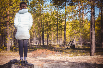 Girl in light jacket is standing on stump and looking deep into pine forest.