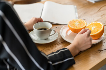 Photographed over the shoulder of a woman, sits at a healthy breakfast with coffee and two orange halves and a fashion magazine