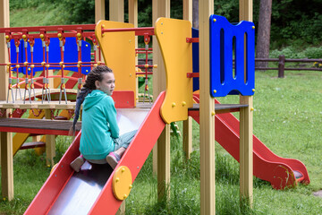 Girl with dreadlocks on her head rides on playground with slide.
