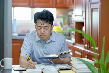 Asian middle-aged man sitting at desk at home, reading a book and studying.