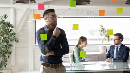 Focused Arabian male employee brainstorm engaged in creative thinking use sticky notes on glass office wall, pensive Arabic businessman develop company business plan or project with post it stickers