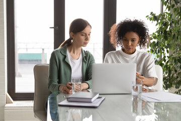 Concentrated multiracial women workers cooperate work together on laptop at office briefing, focused multiethnic female colleagues look at laptop screen brainstorm at meeting, teamwork concept