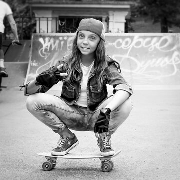 Nice girl, teenager, in a baseball cap sits on a skateboard in a skate park. Black and white photo. Youth Lifestyle. Sport.