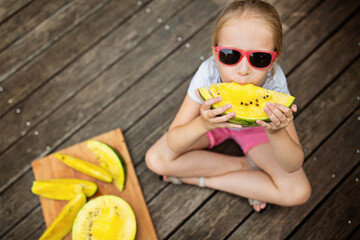 Happy blonde little girl sitting outdoor on wooden bridge and eating yellow watermelon. Top view, overhead shot. Summer local vacation 2020 during coronavirus covid-19 pandemic quarantine.