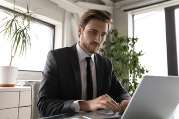 Concentrated Caucasian male employee sit at desk in office work on laptop consult client or customer online, focused young businessman busy using computer internet connection, technology concept