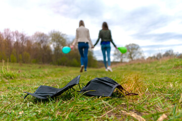 Two girls, holding hands and holding a balloon in their hands, go away from the discarded black medical masks that lie on the grass in the foreground. The girls in the background are blurry. 